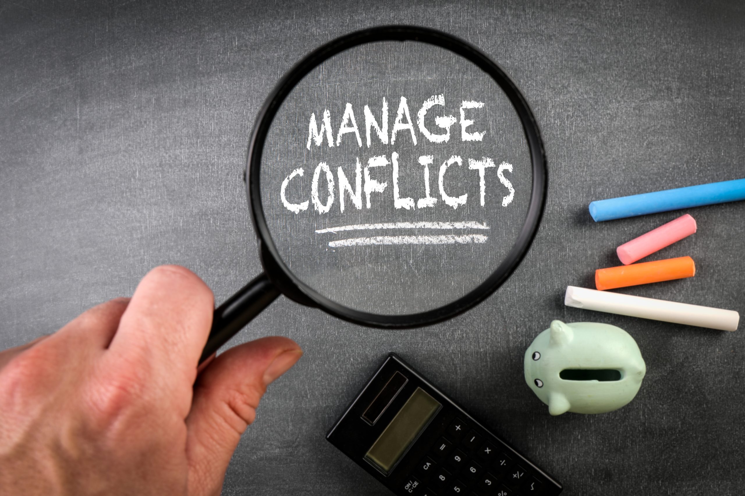 "MANAGE CONFLICTS" écrit sous une loupe avec des craies, une tirelire et une calculatrice