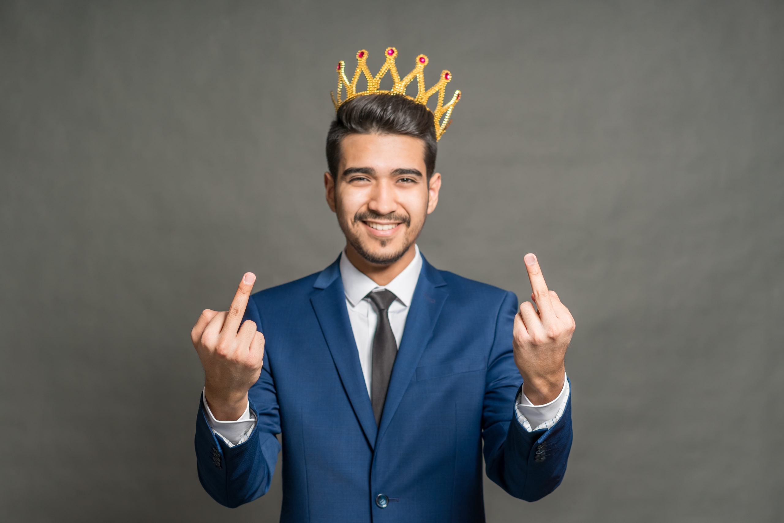 Un homme en costume avec une couronne dorée sourit tout en levant les deux doigts du milieu