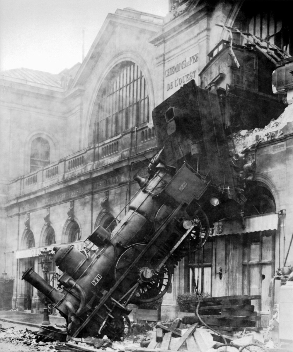 L'image historique montre un accident spectaculaire de train survenu à la gare Montparnasse à Paris en 1895. Une locomotive à vapeur, inclinée à la verticale, a défoncé le mur de la gare et s'est écrasée dans la rue en contrebas. La façade de la gare, portant l'inscription "Chemin de fer de l'Ouest", est visible en arrière-plan, avec des dégâts importants autour de la zone impactée. Cette scène illustre un événement marquant et rare dans l'histoire ferroviaire.