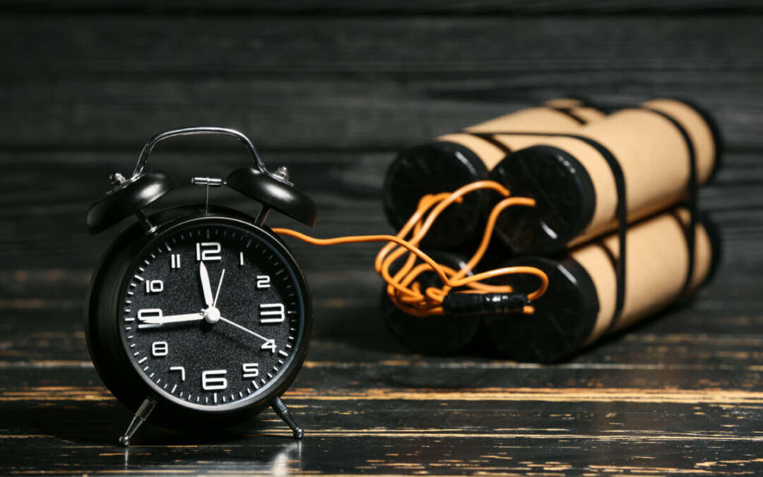 L'image montre un réveil noir classique posé sur une surface en bois sombre, relié par des fils électriques à plusieurs cylindres enroulés de bande noire, rappelant un dispositif explosif. Cette scène évoque la notion de "bombe à retardement", symbolisant une situation ou une tension qui pourrait éclater à tout moment si elle n'est pas désamorcée.