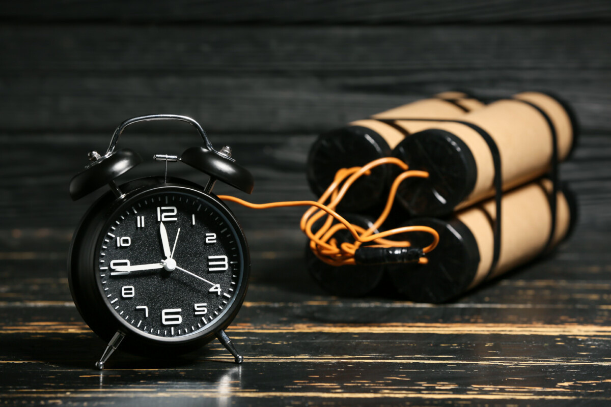 L'image montre un réveil noir classique posé sur une surface en bois sombre, relié par des fils électriques à plusieurs cylindres enroulés de bande noire, rappelant un dispositif explosif. Cette scène évoque la notion de "bombe à retardement", symbolisant une situation ou une tension qui pourrait éclater à tout moment si elle n'est pas désamorcée.