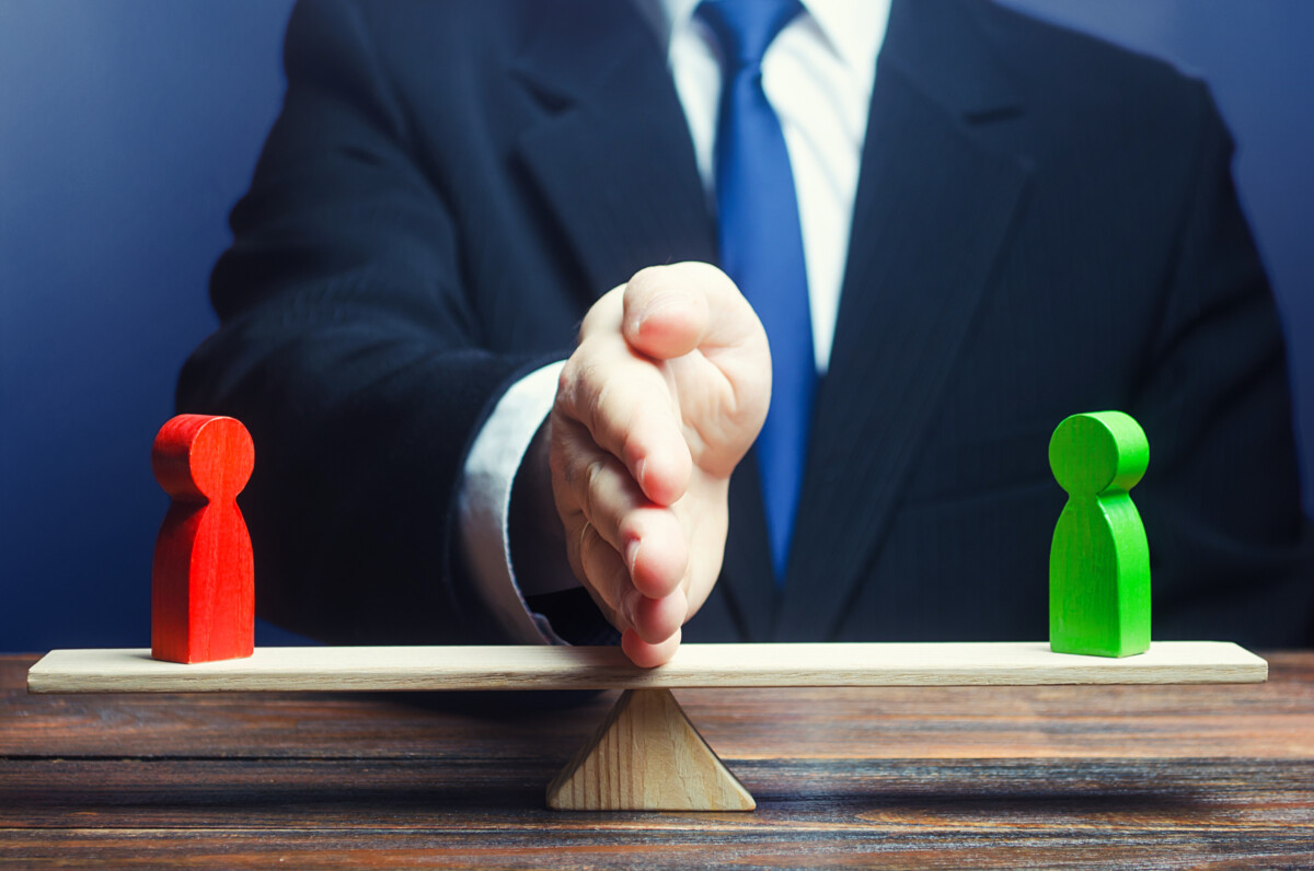 Un médiateur, représenté par une main tendue, sépare deux figurines en bois, l'une rouge et l'autre verte, posées sur une balance en équilibre. L'image symbolise la médiation ou l'intervention pour résoudre un conflit entre deux parties opposées, illustrant l'équilibre, la neutralité et la recherche d'une solution équitable.