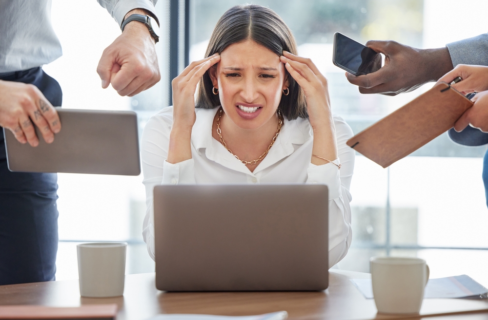 Une femme, assise devant un ordinateur portable, affiche une expression de stress intense. Elle se tient la tête entre les mains tandis que plusieurs mains autour d'elle lui tendent des tablettes, un téléphone et des documents, symbolisant une surcharge de travail ou des demandes pressantes dans un contexte professionnel.