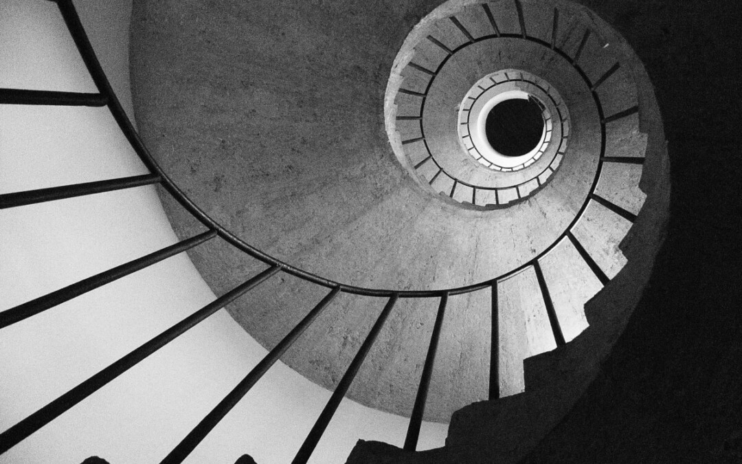 Un escalier en colimaçon vu en contre-plongée, capturé en noir et blanc. Les lignes des marches et de la rampe convergent vers un point central lumineux, créant une illusion visuelle de profondeur et de mouvement circulaire. L'image transmet une impression de géométrie, de symétrie et d'infini. Oeuvre de Vincenzo Elviretti