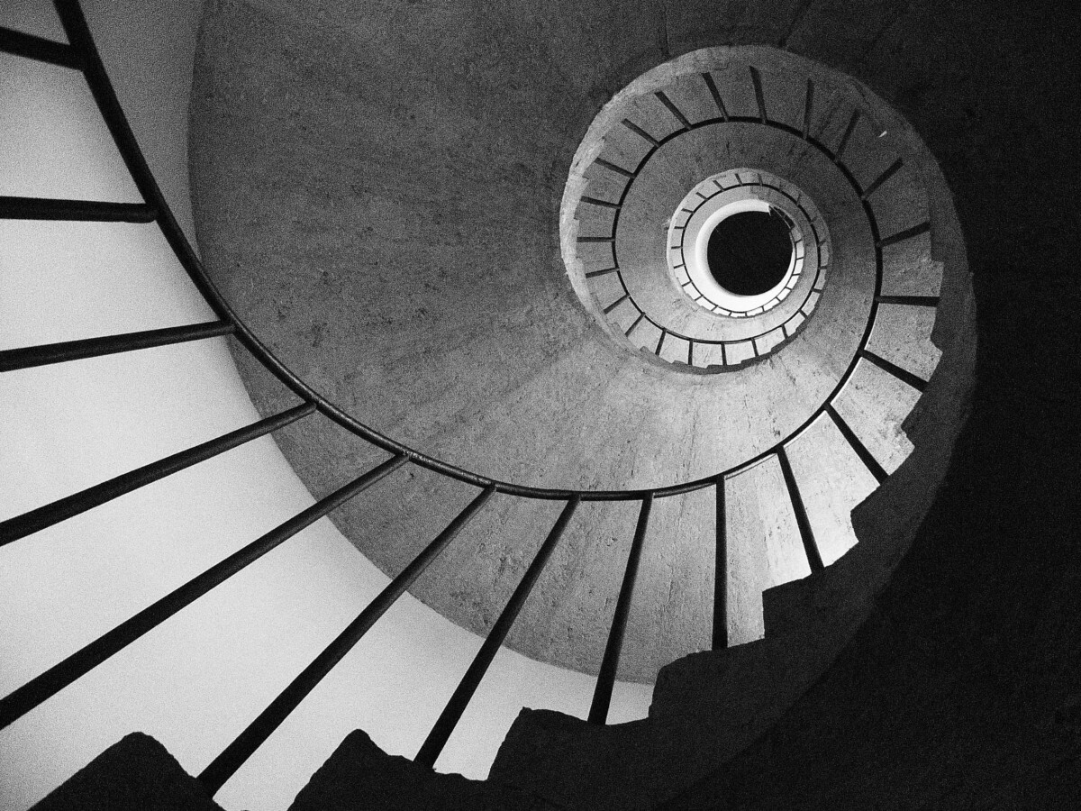 Un escalier en colimaçon vu en contre-plongée, capturé en noir et blanc. Les lignes des marches et de la rampe convergent vers un point central lumineux, créant une illusion visuelle de profondeur et de mouvement circulaire. L'image transmet une impression de géométrie, de symétrie et d'infini. Oeuvre de Vincenzo Elviretti