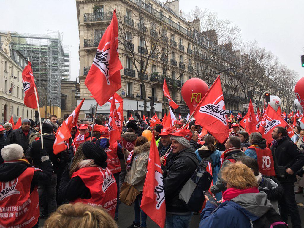 Une manifestation rassemblant un grand nombre de personnes dans une rue urbaine, brandissant des drapeaux rouges et des ballons aux couleurs de l'organisation syndicale "FO" (Force Ouvrière). Les manifestants portent des gilets rouges, et l'ambiance reflète une mobilisation collective dans un contexte revendicatif.