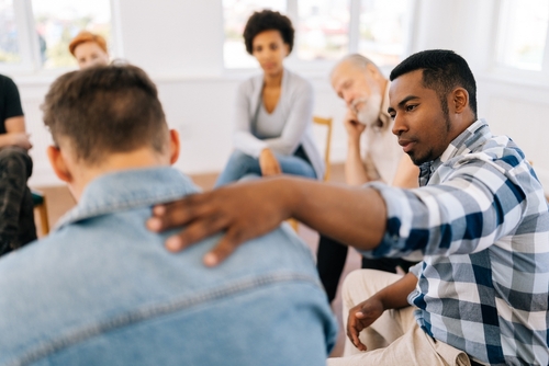 Un groupe de personnes assises en cercle, engagé dans une discussion ou une séance de soutien. Un homme pose une main réconfortante sur l'épaule d'un autre participant, symbolisant l'empathie, le soutien mutuel et la compréhension dans un environnement bienveillant. L'arrière-plan lumineux suggère une ambiance sereine et propice à l'écoute.