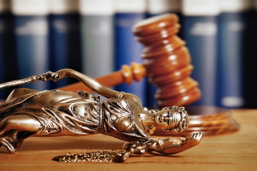 Une statue miniature de la déesse de la justice, Lady Justice, est renversée sur une table en bois, ses balances et son épée à côté d'elle. En arrière-plan, un maillet en bois, symbole du pouvoir judiciaire, est flou, avec des rangées de livres de droit visibles derrière. Cette composition semble évoquer des notions de déséquilibre ou de remise en question de la justice.