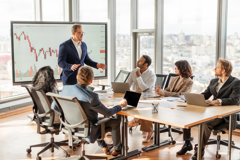 Un groupe de professionnels en réunion dans une salle de conférence moderne avec une vue panoramique sur la ville. Un homme en costume bleu, debout devant un écran affichant un graphique financier, semble expliquer une tendance à ses collègues assis autour de la table. Ces derniers, équipés d'ordinateurs portables et de documents, l'écoutent avec attention. L'ambiance est sérieuse et studieuse, illustrant une analyse de marché ou une prise de décision stratégique en entreprise.